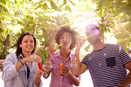 Three funloving people laughing and blowing bubbles in the dappled afternoon sunshine with some trees around them wearing casual clothingの写真素材