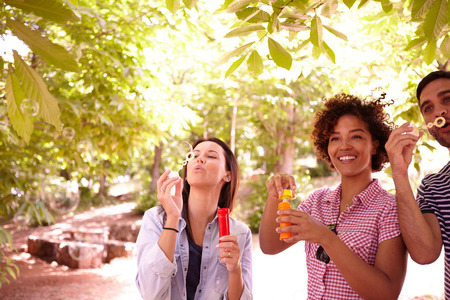 Three young friends laughing and blowing bubbles in the dappled afternoon sunshine with some trees around them wearing casual clothingの写真素材