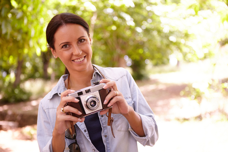 Smiling young woman holding an old style camera in the shade of some trees with dappled sunshine wearing casual clothingの写真素材