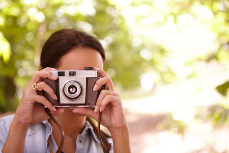 Young female taking a picture with an old style camera in the shade of some trees wearing casual clothingの写真素材