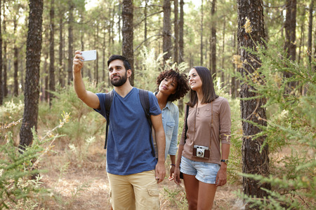Three friends hiking through a pine tree forest in the late afternoon shadows and taking selfies in front of a treeの写真素材