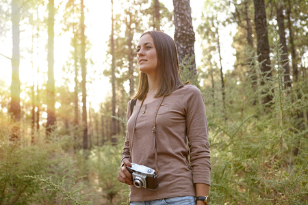 Young girl in a forest of pine trees holding her camera wonderingly looking ahead of herself while wearing a brown t-shirt and shortsの写真素材