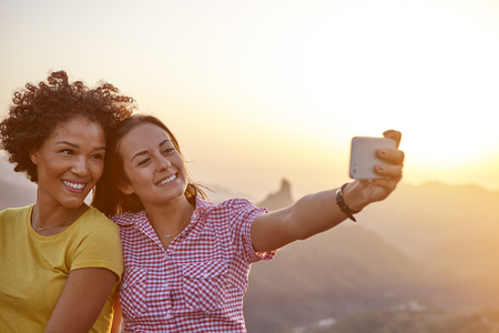 Two girl friends taking a selfie with mountains behind them while sitting on a stone wall and wearing casual clothingの写真素材