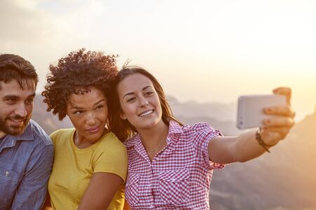 Three friends taking a selfie with mountains behind them while sitting on a stone wall and wearing casual clothingの写真素材