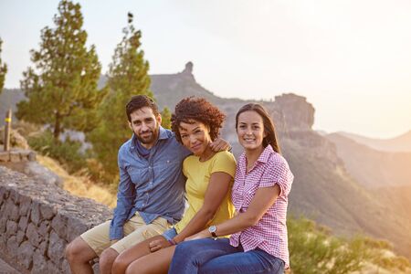 Three friends posing for a picture in the late afternoon sunshine sitting on a stone wall while wearing casual clothingの写真素材