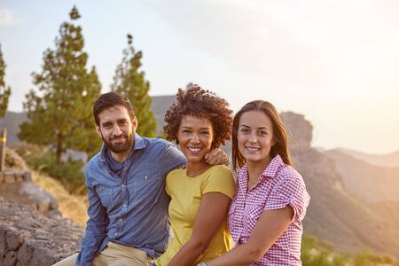 Three friends posing for a picture with mountains behind them, sitting on a stone wall and wearing casual clothingの写真素材