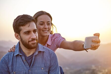 Smiling young couple taking a selfie on a mountain top with mountains behind them and wearing casual clothingの写真素材
