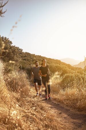 Couple running down a dusty path in full sun light in full sun light with bushes behind them while wearing black and greyの写真素材