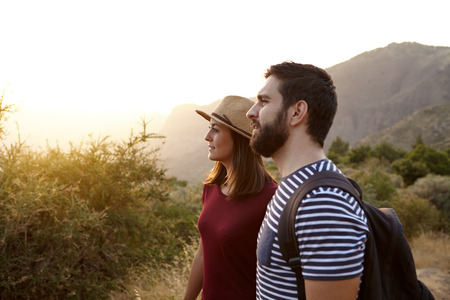 Young couple looking at something far away surrounded by bushes and very bright sun to their right wearing t-shirts and a straw hat at sunsetの写真素材