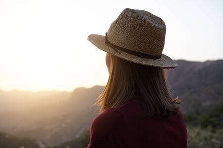 Girl from behind looking at something over the mountains in very bright sun in front of her wearing...