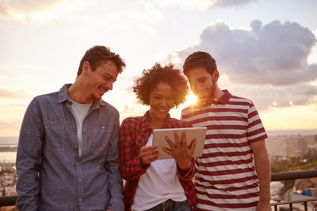 Three happy friends looking at the tablet joyously while the sun is setting behind them, wearing casual clothingの写真素材