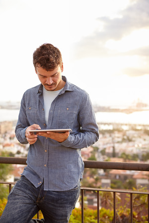 Happy young man on a bridge that overlooks the city, typing on his tablet while wearing casual clothingの写真素材
