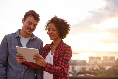 Smiling couple looking at a tablet with interest while the sun is setting behind them, wearing casual clothingの写真素材