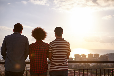 Three friends on a bridge looking out over the city at a sun setting while wearing casual clothing their hands in front of themの写真素材