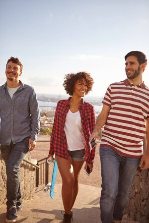 Three joking friends with a skateboard and cell phone laughing while walking up steps with big smiles and pale blue skies behind themの写真素材