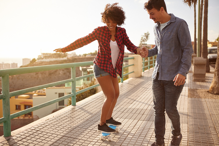 Boyfriend showing girl how to skateboard while holding her hand tightly and walking her slowly while she has her other arm outの写真素材