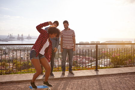 Three cool friends with a skateboard on a bridge, all laughing while the girl is trying to keep her balance on the skateboardの写真素材