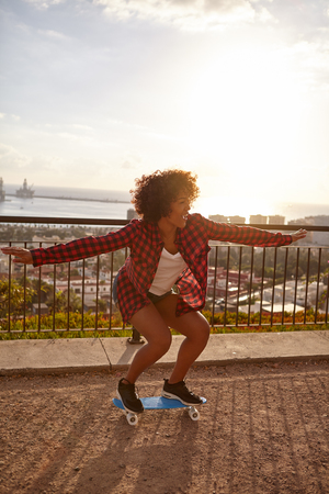 Skateboarding girl with her arms out and knees bent, on a bridge overlooking a city with a bay further behind, wearing casual clothingの写真素材