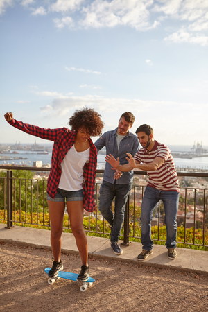 Three young friends playing with a skateboard on a bridge, the men holding their hands out to catch the girl if she fallsの写真素材