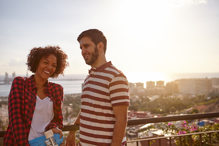 Laughing cool couple on a bridge with a metal railing overlooking a city and bay with bright sunshine to the rightの写真素材