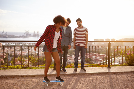 Two male friends on a bridge above a city watching a girl balancing on a skateboard, all wearing casual clothingの写真素材
