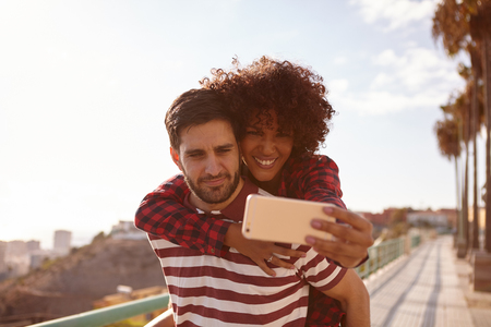 Cute young couple taking playful selfies with her sitting on her back, right arm around his neck, a toothy smile and him holding her upの写真素材