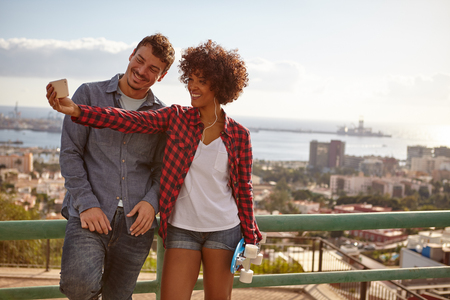 Laughing couple posing with cell phone while holding a blue skateboard and laughing as they lean against metal railing of a bridgeの写真素材