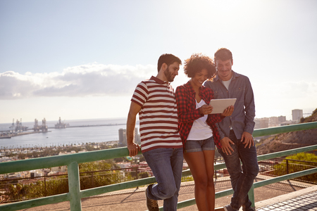 Three friends leaning against a railing, smiling at the tablet, the girl is holding while standing between the young menの写真素材