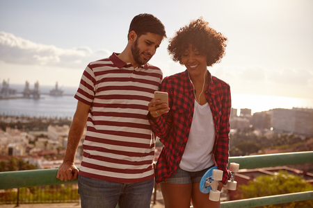 Laughing couple looking at cell phone while holding a blue skateboard and leaning on the metal railing with a cityscape behind themの写真素材
