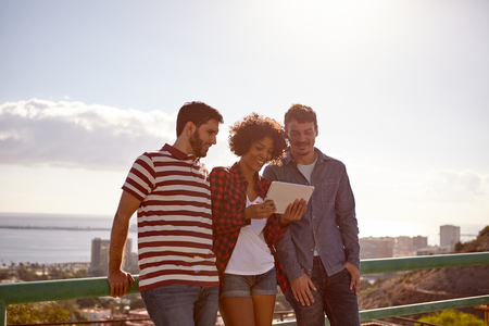 Three friends smiling at a tablet with very bright late afternoon sun behind them and a city scape and bay areaの写真素材
