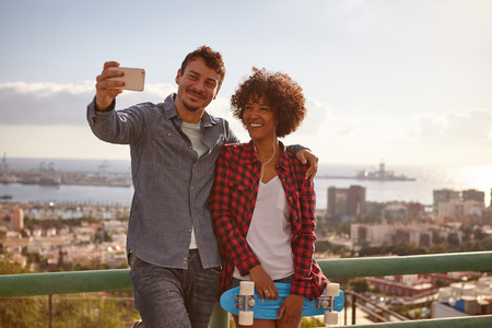 Smiling couple posing with cell phone while she holds a blue skateboard and laughing as they lean...