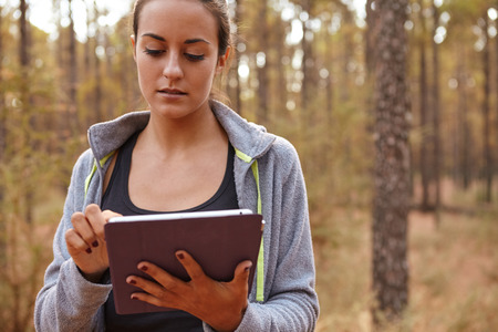 Sweet young girl concentrating on her tablet while holding it with her left hand and using her right index finger while in a forestの写真素材