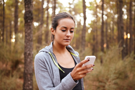 Concentrating young girl with her cellphone looking at her cellphone while standing alone in the forest in natural daylightの写真素材