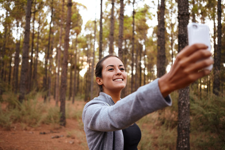 Lovely smiling young girl taking a selfie with her cell phone while surrounded by trees in a forest with natural sunlightの写真素材