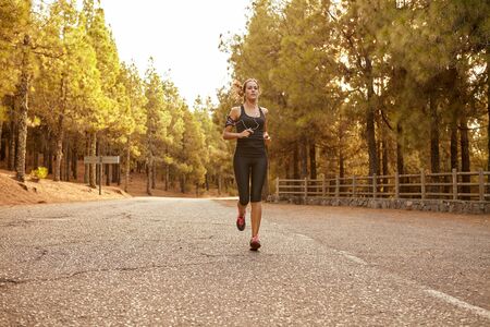 Young brunette woman running in a forest on a tarred road cutting through it with bright sunshine from behind herの写真素材