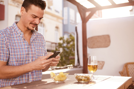 Handsome young man with short dark hair looking at his cellphone while sitting at a table with a drink and snacksの写真素材