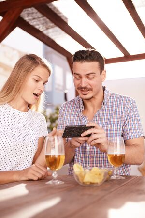 Surprised blond looking at something on the cellphone he is explaining to her with a quirky smile and pointing at itの写真素材