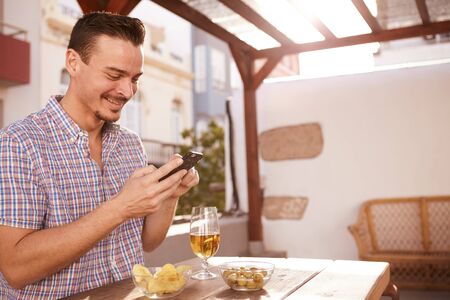 Goodlooking young man looking at his cell with a happy smile while sitting at a table with a drink and snacksの写真素材