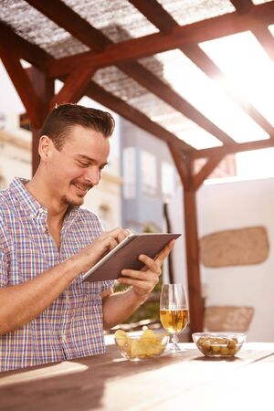 Dark and handsome young guy busy with his tablet while sitting at a table with a drink and bowls of snacksの写真素材