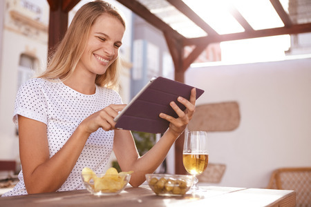 Pretty blond haired girl pointing at something on her tablet with a toothy smile while sitting at a table with a drinkの写真素材