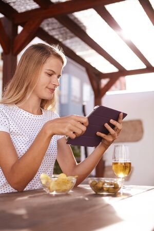 Beautiful blond haired girl looking at her tablet in deep concentration while sitting at a table with a drink and snacksの写真素材