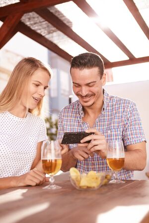 Happy couple looking at a cellphone with big smiles while sitting at a table with drinks snacks in very bright sunlight from behindの写真素材