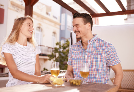 Laughing young couple having drinks in front of a big window while sitting at a wooden table with bowls of snacksの写真素材