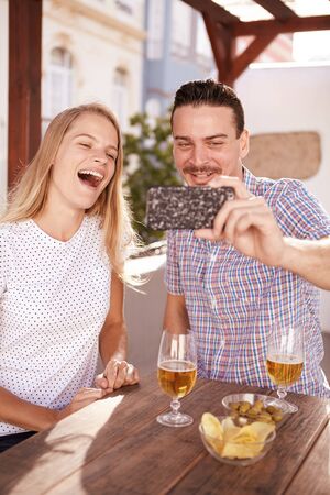 Laughing couple taking a happy selfie while sitting at a wooden table with drinks and bowls of snacks in bright sunlightの写真素材