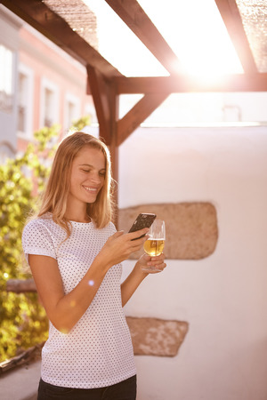 Lovely blond girl with a toothy smile looking at her celphone with a beer in the other hand and bright sunshine from behindの写真素材