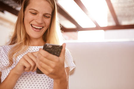 Happily laughing blond girl with her cellphone in one hand and touching the phone with her other index fingerの写真素材