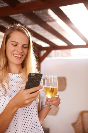 Happily smiling blond girl using her cellphone while holding a beer in the other hand while her long hair is hanging looseの写真素材