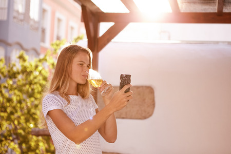 Lovely blond girl sipping on a beer while looking at her cellphone with bright over exposed sunshine behind herの写真素材
