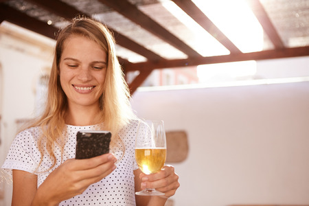 Very happily smiling cute blond woman using her cellphone while holding a beer in the other hands and her long hair hanging looseの写真素材