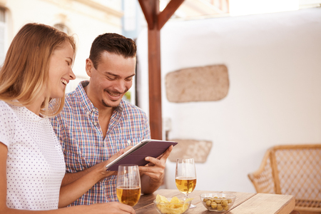 Happy young couple laughing while sharing a touchpad relaxing with beers and snacks on the table and bright sunshine behind themの写真素材